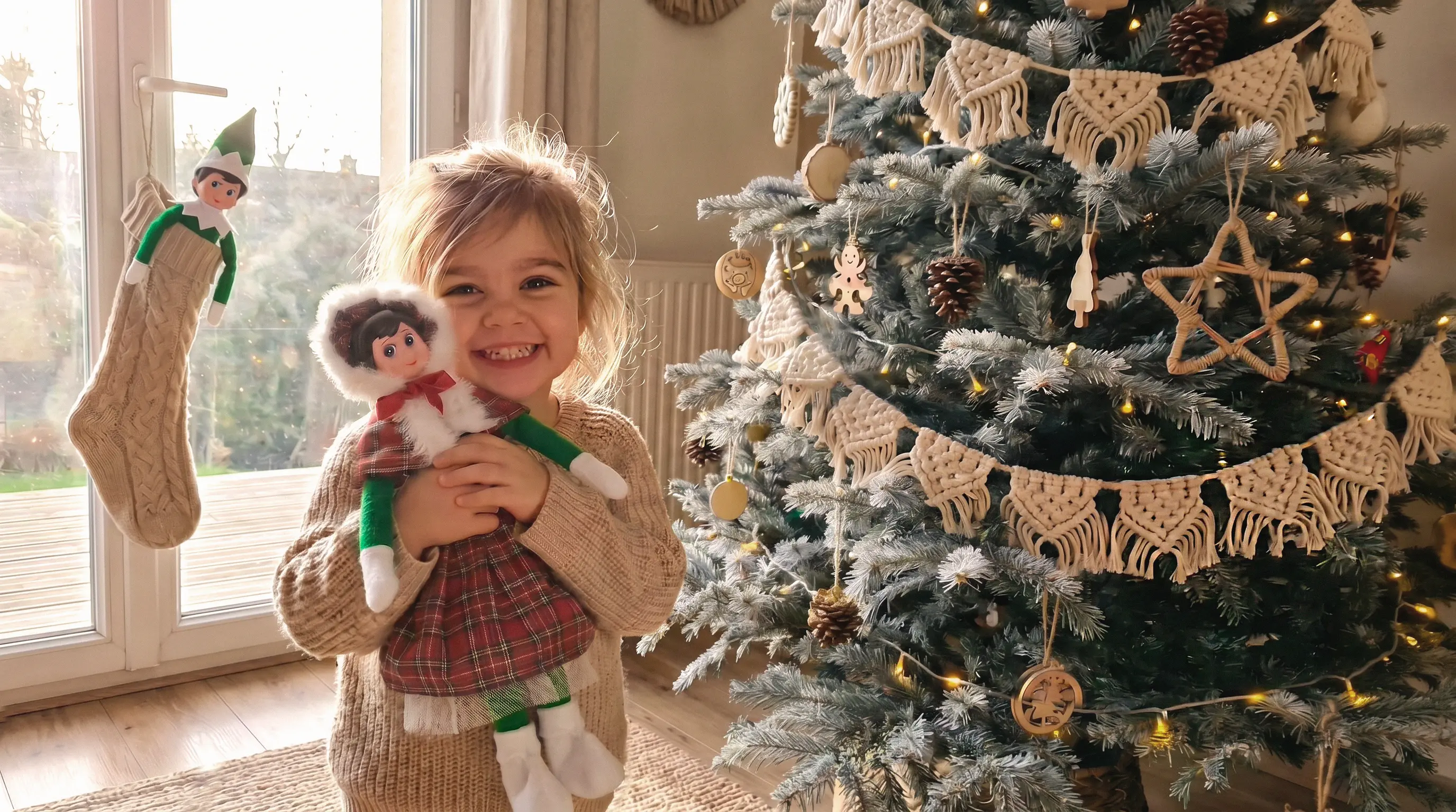 Une petite fille tout sourire serre son Lutin Farceur devant le sapin décoré